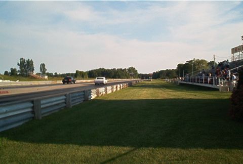 1985 Ford LTD Crown Victoria - A drag race is underway on a sunny day with a crowd watching from the bleachers.
