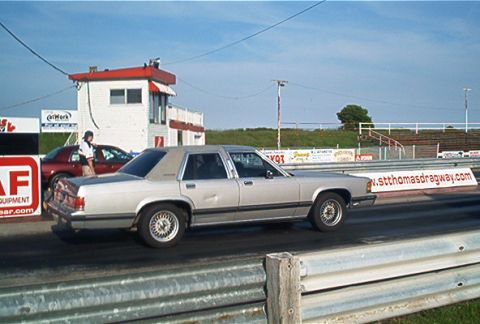 1985 Ford LTD Crown Victoria - A silver sedan is on the drag strip, with another car and a building in the background.