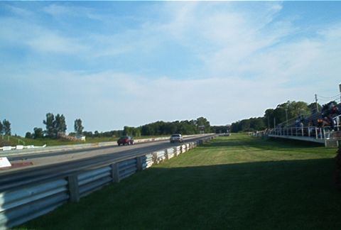 1985 Ford LTD Crown Victoria - A drag race is underway with two cars on the track and spectators watching from the stands.