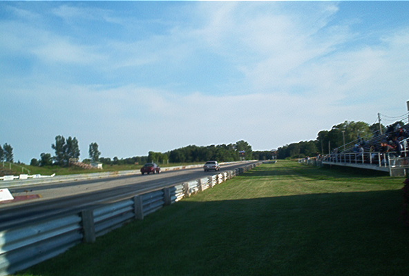 Two cars racing on a drag strip with spectators in bleachers