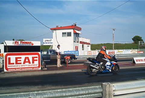 1985 Ford LTD Crown Victoria - Motorcycle races down a drag strip past a pickup truck and a timing tower.