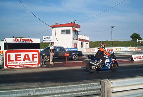 1985 Ford LTD Crown Victoria - A motorcycle and a car are staged at a drag strip, with a control tower and advertising signs visible.