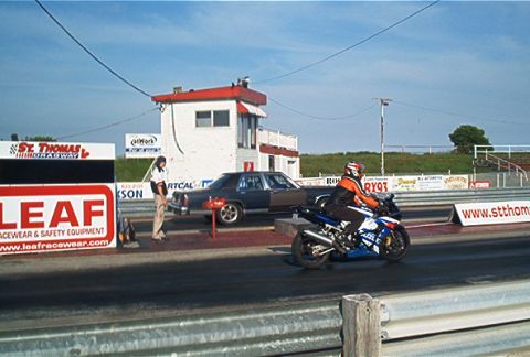 1985 Ford LTD Crown Victoria - A dark sedan and a blue motorcycle are positioned on a drag strip, with a person standing near the sedan.