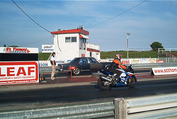 Dark sedan and blue motorcycle on a drag strip, with a person standing by the car