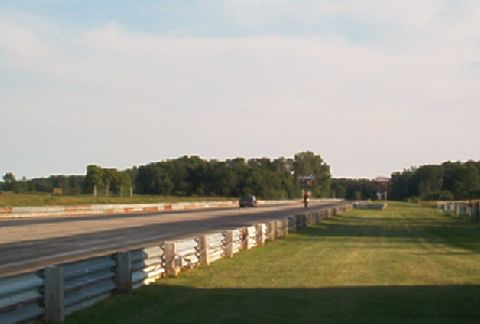 1985 Ford LTD Crown Victoria - A lone car travels down a drag strip, bordered by grass and trees under a bright sky.