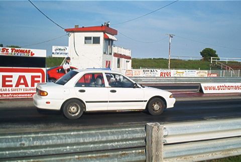 1985 Ford LTD Crown Victoria - A white sedan is on the racetrack, with a red car and a person visible in the background near a tower.