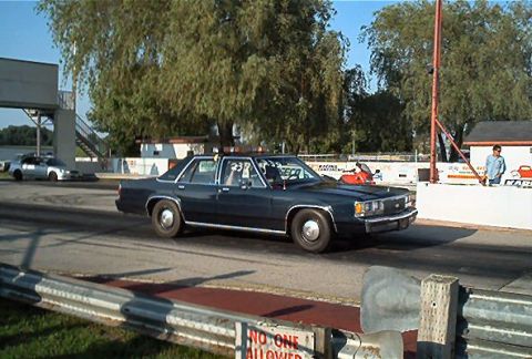 1985 Ford LTD Crown Victoria - A dark blue sedan with a roll cage is on a race track, with another car in the background.