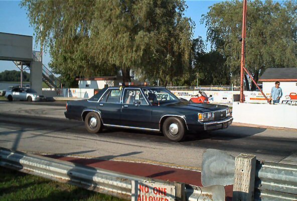 Dark blue sedan with chrome trim, driving on a track. Visible roll cage inside. Another silver sedan is in the background