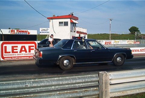 1985 Ford LTD Crown Victoria - A dark blue 1985 Ford Crown Victoria LTD police car is seen at a drag strip, with other vehicles and signage in the background.