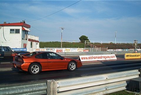 1985 Ford LTD Crown Victoria - A drag race is underway with an orange sports car in the foreground and a building in the background.