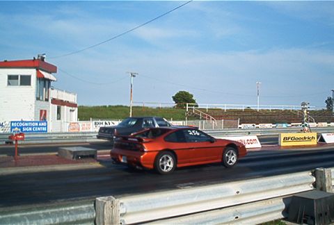 1985 Ford LTD Crown Victoria - Two cars compete on a drag strip under a clear blue sky, with spectators' seating and a building visible.