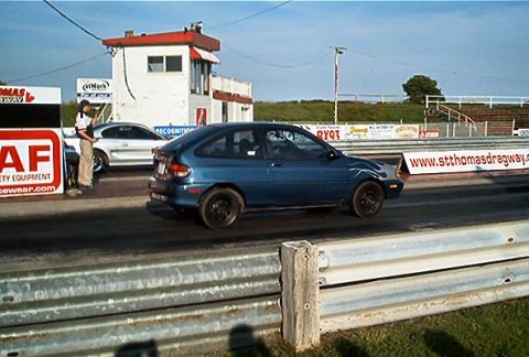 1985 Ford LTD Crown Victoria - A blue hatchback competes on a drag strip, with a silver car and a person visible in the background.