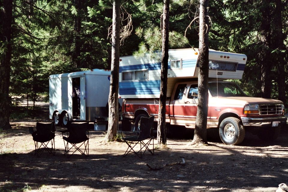 Red Ford pickup with slide-in camper towing enclosed trailer at forest campsite