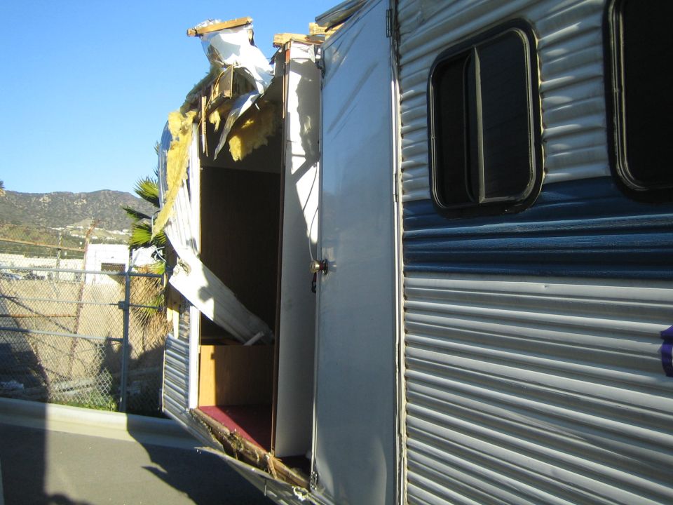 Damaged trailer with roof torn off and insulation exposed, side door open, blue stripe on exterior