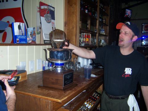 1985 Ford LTD Crown Victoria - A bartender pours a drink into a trophy at a bar, with a football poster in the background.