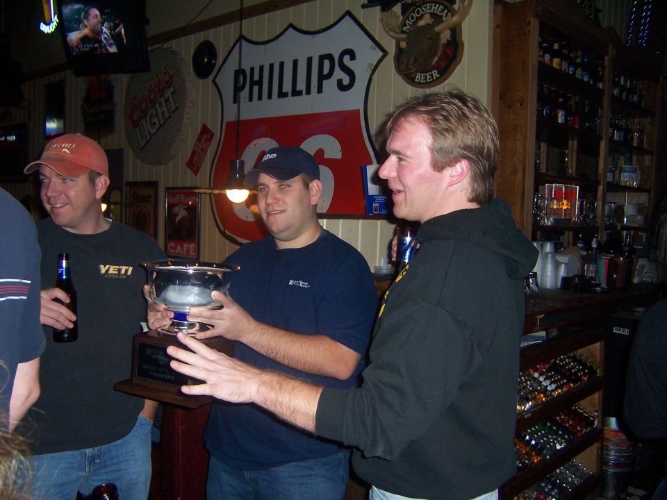 Three men in a bar, one holding a trophy, with Route 66 and Phillips 66 signs on the wall