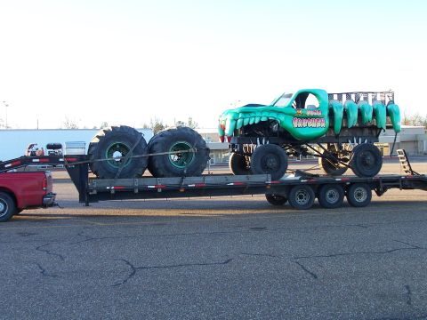 1982 Ford Bronco - A green monster truck named "CRUSHER" is transported on a flatbed trailer, towed by a red pickup truck.