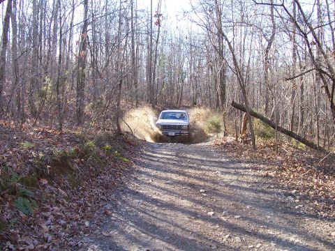 1982 Ford Bronco - A white pickup truck splashes through a muddy puddle on a wooded dirt road.