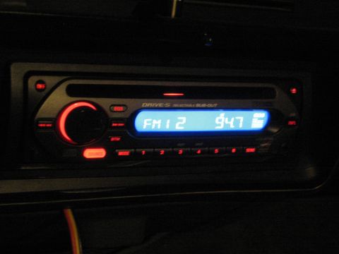 1990 Ford Bronco - Close-up of an aftermarket car stereo faceplate glowing blue, its display reading FM1 2 and 947 with a red volume knob.