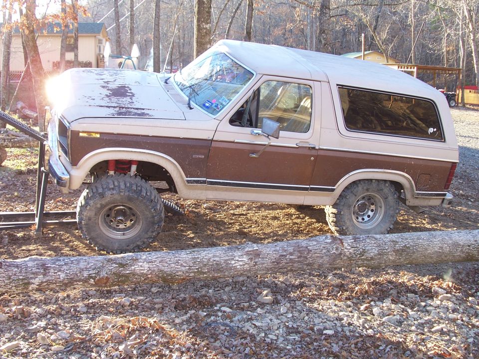 Two-tone brown and tan Ford Bronco with lifted suspension and large tires on a dirt path with a log