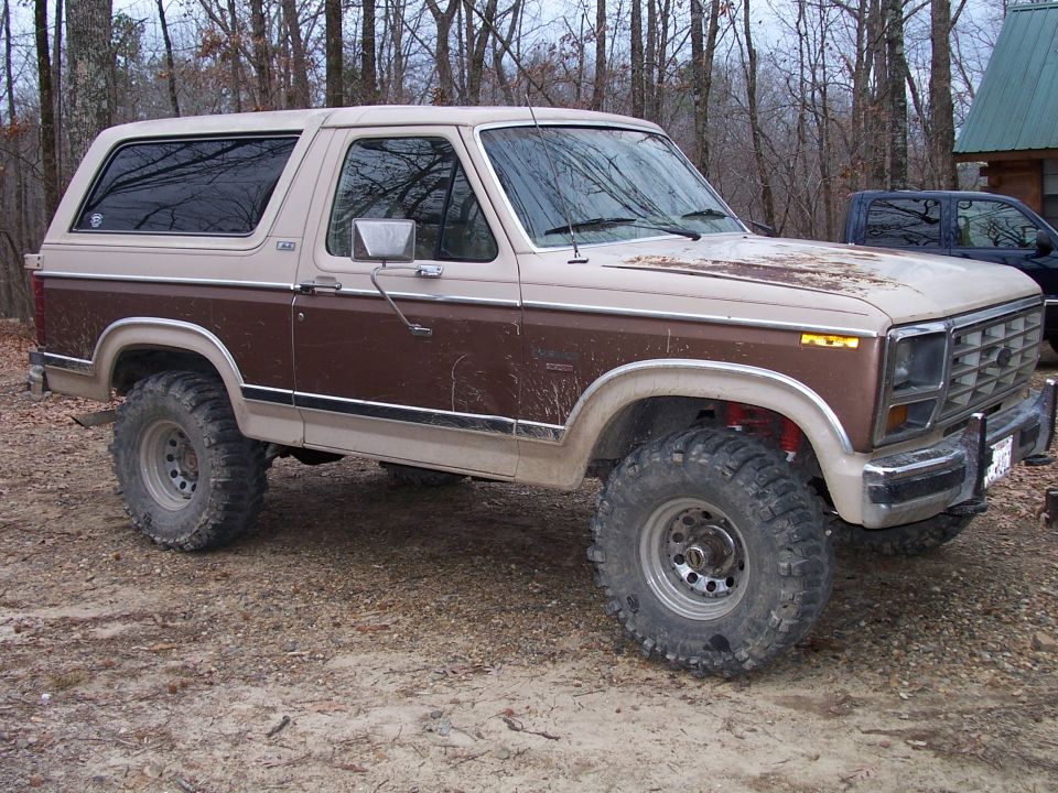 Lifted 1980s Ford Bronco with large off-road tires parked on dirt, another dark pickup truck visible behind it