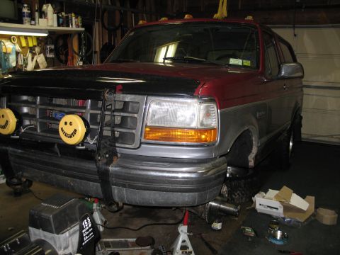1995 Ford Bronco - Front view of a 1995 Ford Bronco in a garage, lifted on jack stands with parts scattered around.