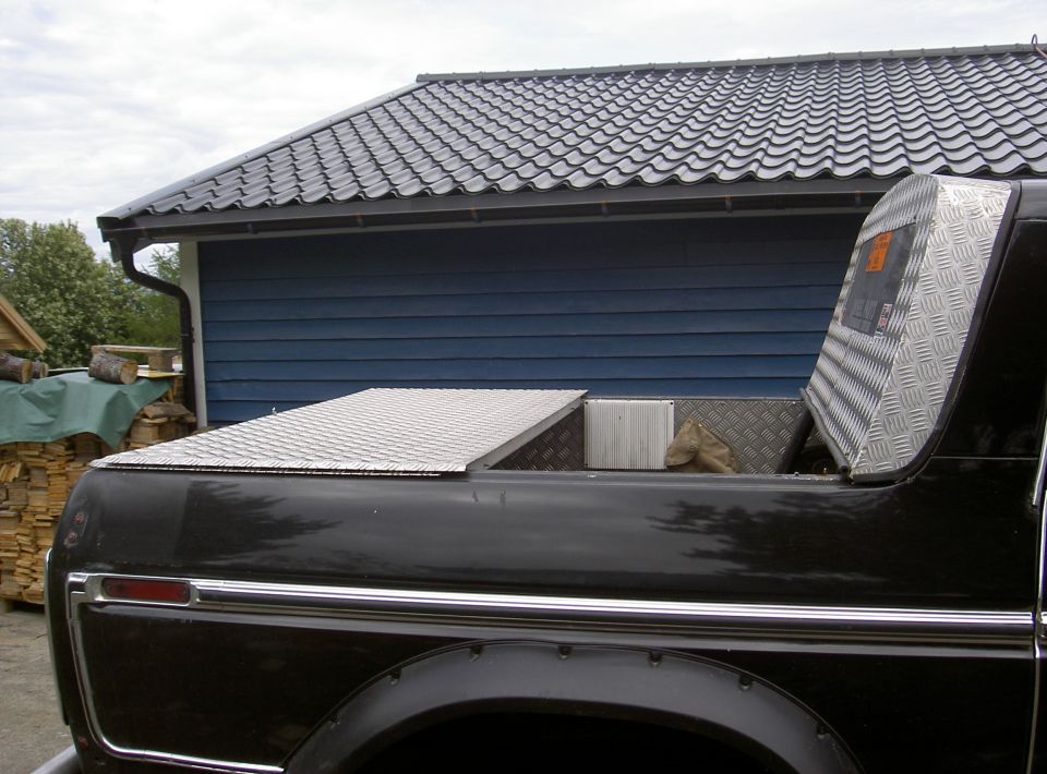 Dark truck bed side view with diamond-plate aluminum cover and bulkhead, blue building behind