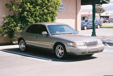2000 Mercury Grand Marquis - A gray 2000 Mercury Grand Marquis is parked in a lot, showing its front three-quarter view with custom chrome wheels.