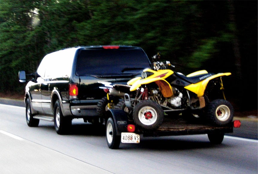 Black SUV towing a trailer with a yellow Honda ATV on it, driving on a road