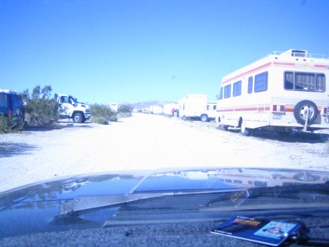 1990 Ford Bronco - Looking over a dark vehicle hood at motorhomes and trucks parked along a dusty desert road under a blue sky.