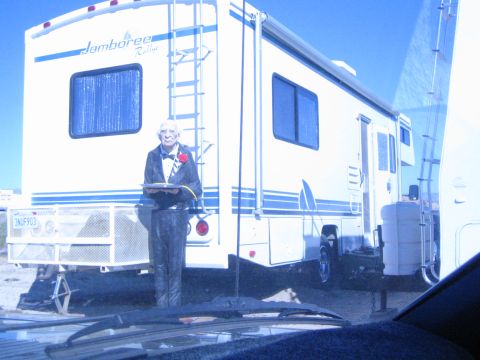 1990 Ford Bronco - person dressed in a tuxedo holding a clipboard and red flower stands beside a white Jamboree motorhome in the desert.
