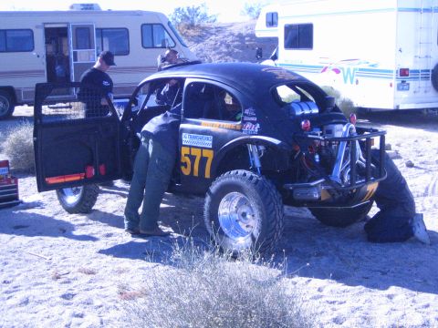 1990 Ford Bronco - black VW Beetle-bodied off-road race car numbered 577 sits in a desert pit area as people work on it beside parked motorhomes.
