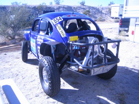 1990 Ford Bronco - blue Volkswagen Beetle-based Baja Bug race car marked 554 sits on knobby tires in a dusty desert race pit.