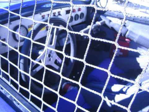 1990 Ford Bronco - Looking through a rope window net at the steering wheel, dashboard and blue interior of an off-road race car cockpit.