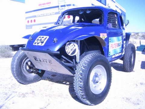 1990 Ford Bronco - blue Baja Bug race car numbered 554 with FilterMag and Elite decals parked on sand beside a trailer.