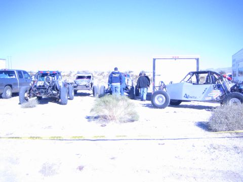 1990 Ford Bronco - Several off-road race buggies and a pickup lined up behind yellow caution tape in a desert staging area as people stand nearby.