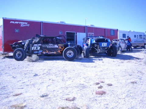 1990 Ford Bronco - black flame-graphic buggy 113 and a blue buggy 131 parked on gravel in front of a red Playtech Racing enclosed trailer.