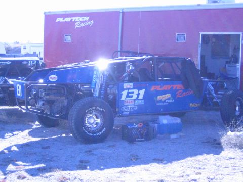 1990 Ford Bronco - blue tube-frame off-road race buggy numbered 131 with Playtech Racing decals parked by a red trailer with a toolbox beside it.