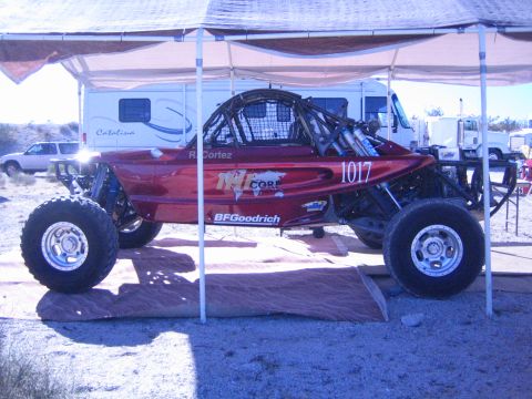 1990 Ford Bronco - Side profile of a dark red tube-frame off-road race buggy numbered 1017 with BFGoodrich decals parked under a canopy near motorhomes.