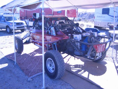 1990 Ford Bronco - Rear three-quarter view of a dark red off-road race buggy 1017 under a canopy, exposing its rear engine and tube frame.