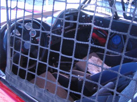 1990 Ford Bronco - View through a black mesh window net into the dark cockpit of an off-road race car, showing gauges, steering wheel and seats.