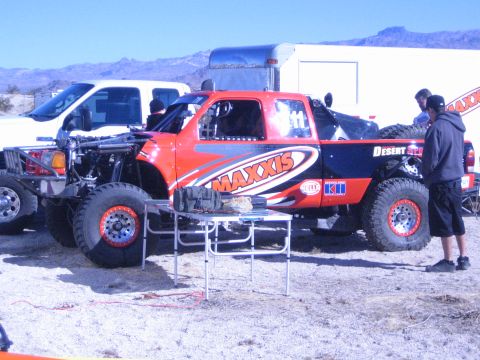 1990 Ford Bronco - orange off-road race truck with Maxxis graphics has its hood open for service as crew work at a folding table in a desert pit.
