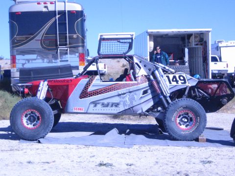 1990 Ford Bronco - silver and red tube-frame off-road race buggy marked 149 with a TYR ram logo parked beside a motorhome in the desert.