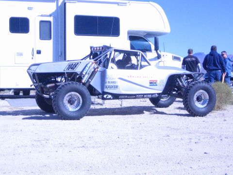 1990 Ford Bronco - silver tube-frame off-road race buggy numbered 1040 sits on knobby tires beside a motorhome as spectators stand nearby.