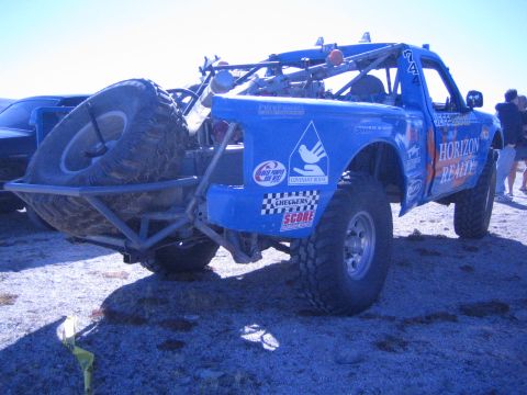 1990 Ford Bronco - blue off-road race truck with Horizon Realty and Score decals carries a spare tire on its bed in a desert pit.