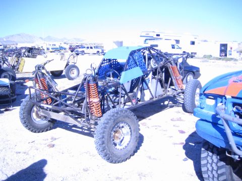 1990 Ford Bronco - bare tube-frame off-road race buggy with orange coilover shocks and no body panels sits among vehicles in a desert pit.