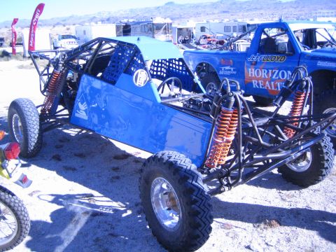 1990 Ford Bronco - blue tube-frame off-road race buggy with orange shocks parked next to a blue Horizon Realty race truck in a desert pit.