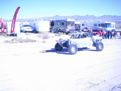 1990 Ford Bronco - white tube-frame off-road race buggy drives across a dusty desert lot lined with motorhomes and trailers.