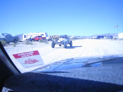 1990 Ford Bronco - View from inside a vehicle through the windshield of an off-road buggy driving ahead on a dusty desert road past parked rigs.