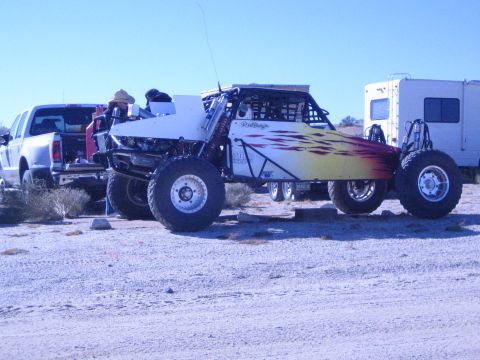 1990 Ford Bronco - white off-road race buggy with yellow-orange flame graphics parked on sand beside a pickup truck and a motorhome.